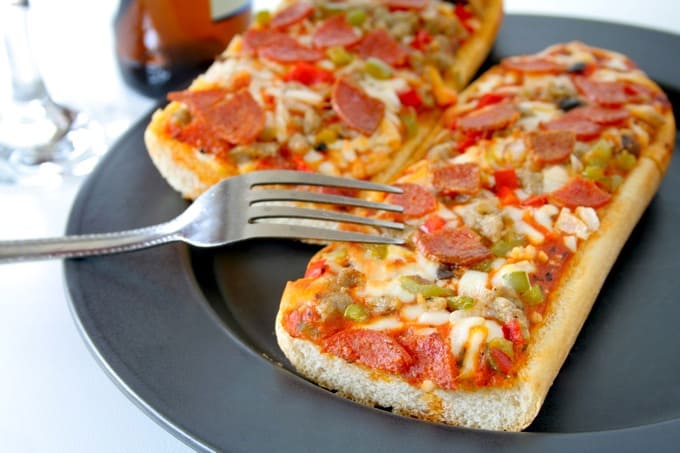 French bread pizza on a dark plate with fork and beverages in the background.