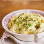 Creamy mashed potatoes in decorative bowl with spoon and garnished with chopped parsley.