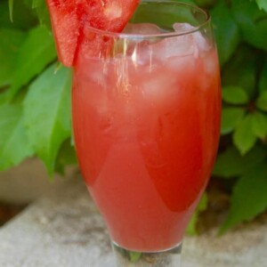 Watermelon Agua Fresca in tall glass with watermelon wedge behind green plant
