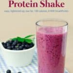 Blueberry peach protein shake in tall glass on table with small bowl of fresh blueberries.