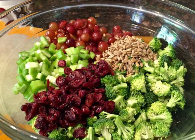 Small broccoli florets, chopped celery, sliced red grapes, dried cranberries and sunflower seeds in large glass bowl