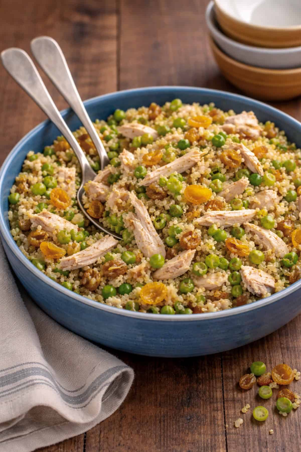 Chicken and Quinoa Salad with peas, raisins and dried apricots in blue bowl with serving spoons on wooden table.