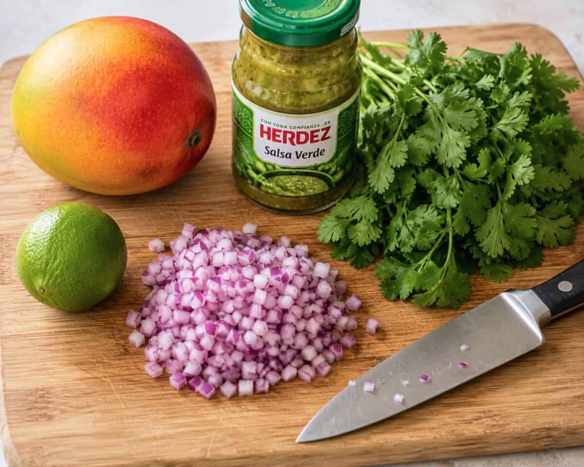Ingredients for making mango salsa, including one whole fresh mango, one lime, a jar of salsa verde, a bunch of cilantro and finely diced red onion on bamboo cutting board with a chef knife.