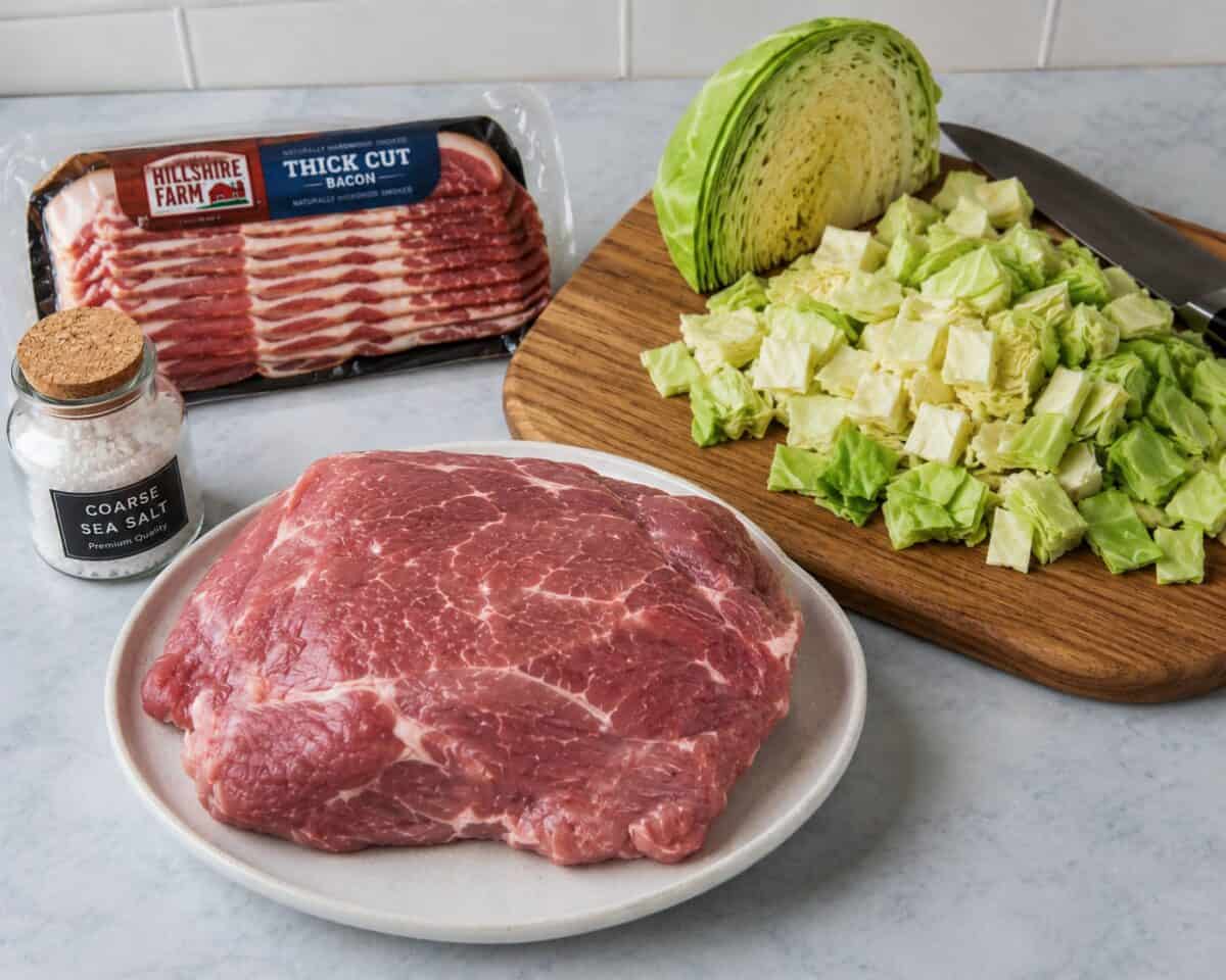 Package of bacon, coarse sea salt, boneless pork shoulder butt roast, and head of green cabbage chopped on wood cutting board with chef knife.