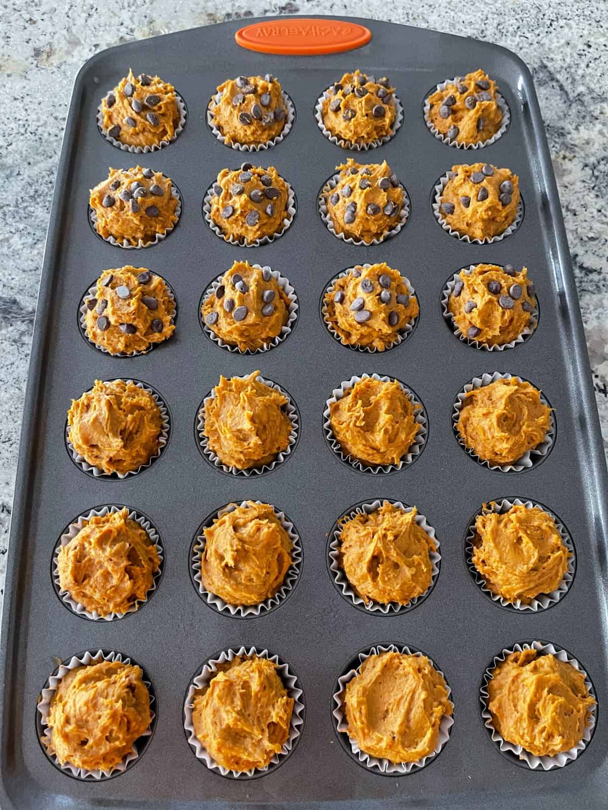 Mini muffin tin with unbaked pumpkin spice cake mix muffins - half plain and half topped with miniature chocolate chips.