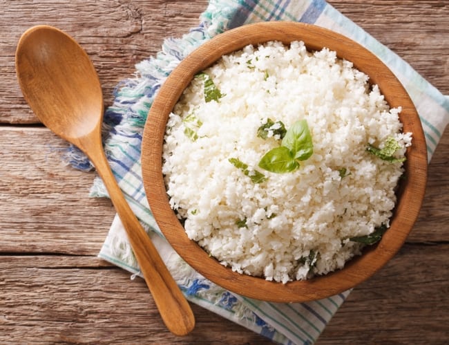 Cauliflower Rice garnished with fresh basil in wooden bowl with wooden spoon.