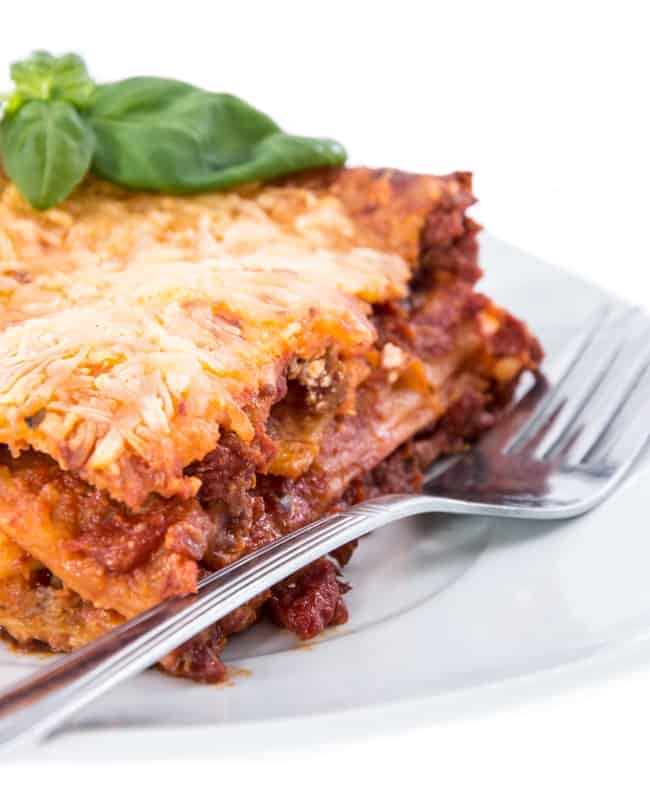 Piece pf lasagna topped with fresh basil on white plate with fork.