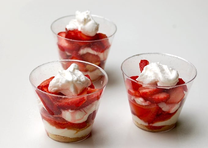 3 clear cups with layered strawberry cheesecake parfaits on white background.