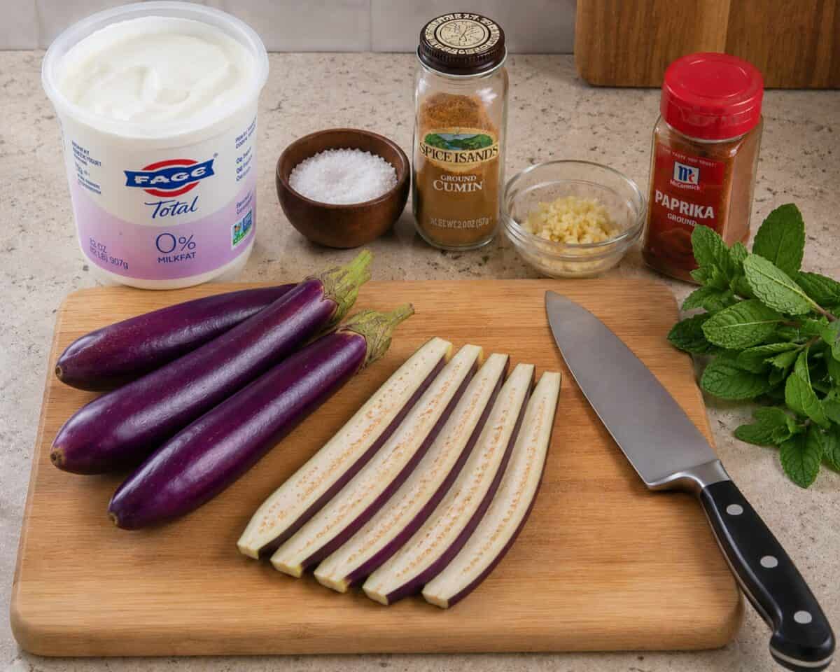 Three Asian eggplant with one eggplant sliced lengthwise on a wood cutting board with chef knife, container of plain, nonfat Greek yogurt, salt, ground cumin, minced garlic, paprika and bunch of mint leaves on kitchen counter.