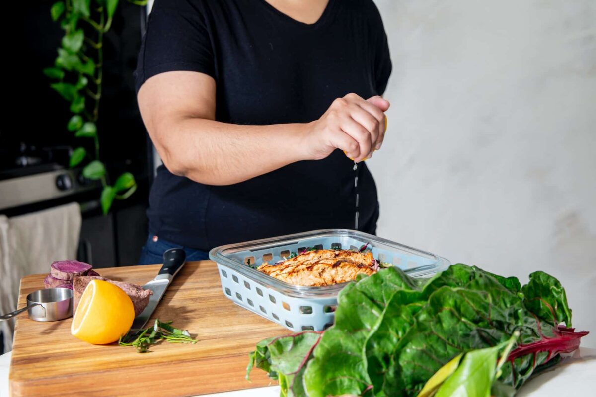 Woman in black tee shirt squeezing lemon over food in a casserole dish with green vegetables in the foreground