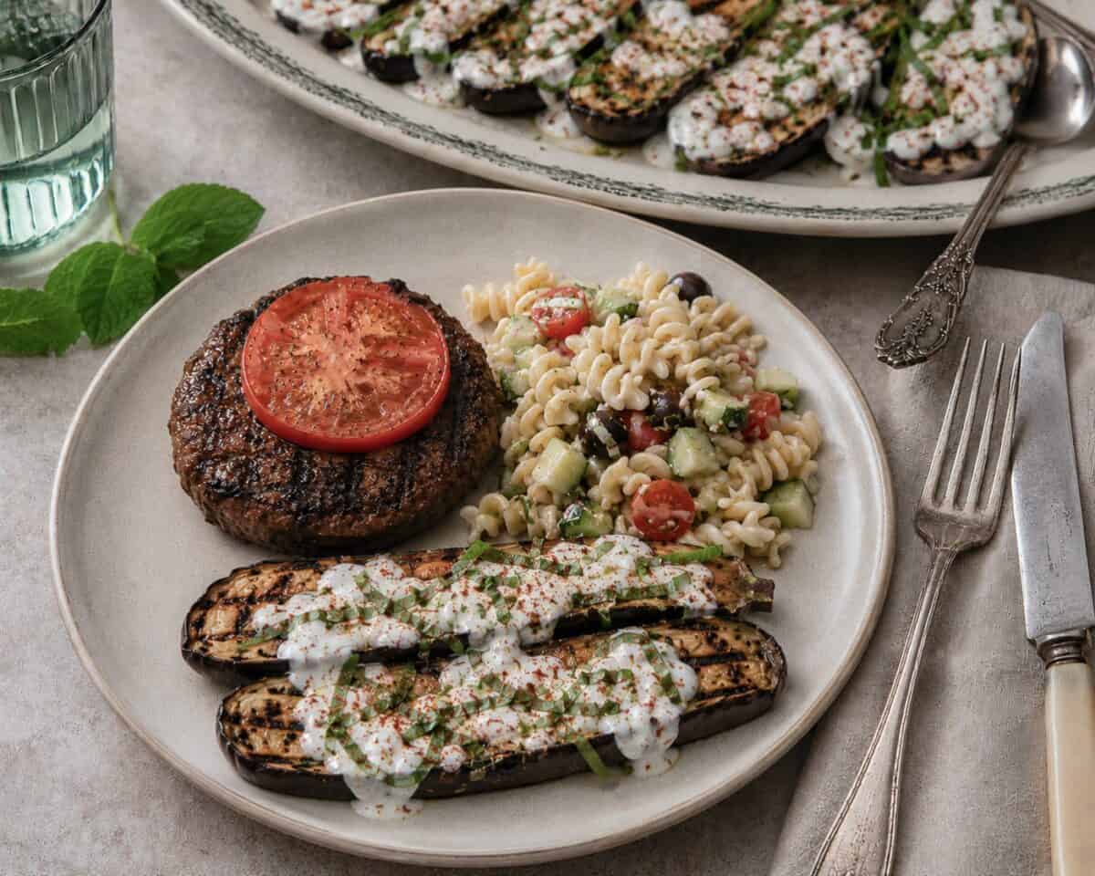 Grilled baby eggplant with yogurt sauce, grilled hamburger with a tomato slice and scoop of pasta salad on dinner plate with platter of grilled eggplant in the background.