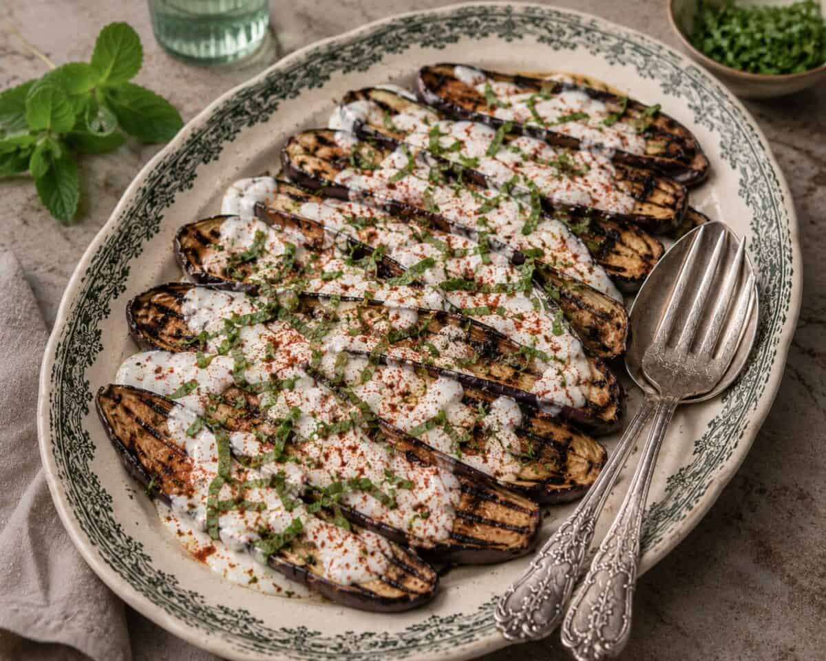 Grilled baby eggplant topped with yogurt sauce, fresh mint and sprinkle of paprika on serving platter with utensils.