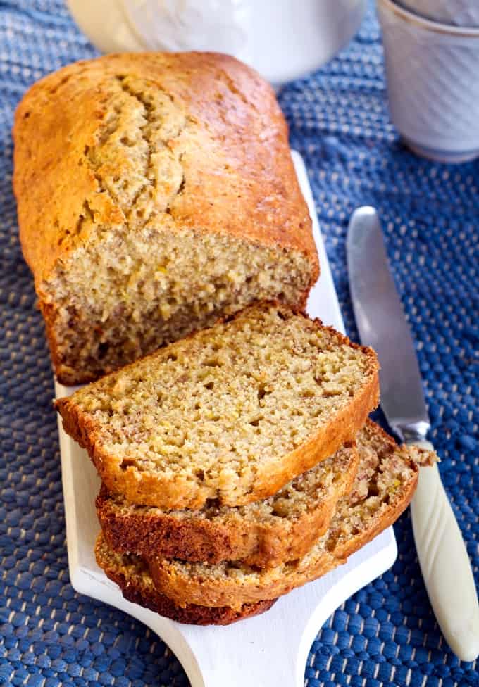 Loaf of banana bread on white cutting board with three slices and knife.