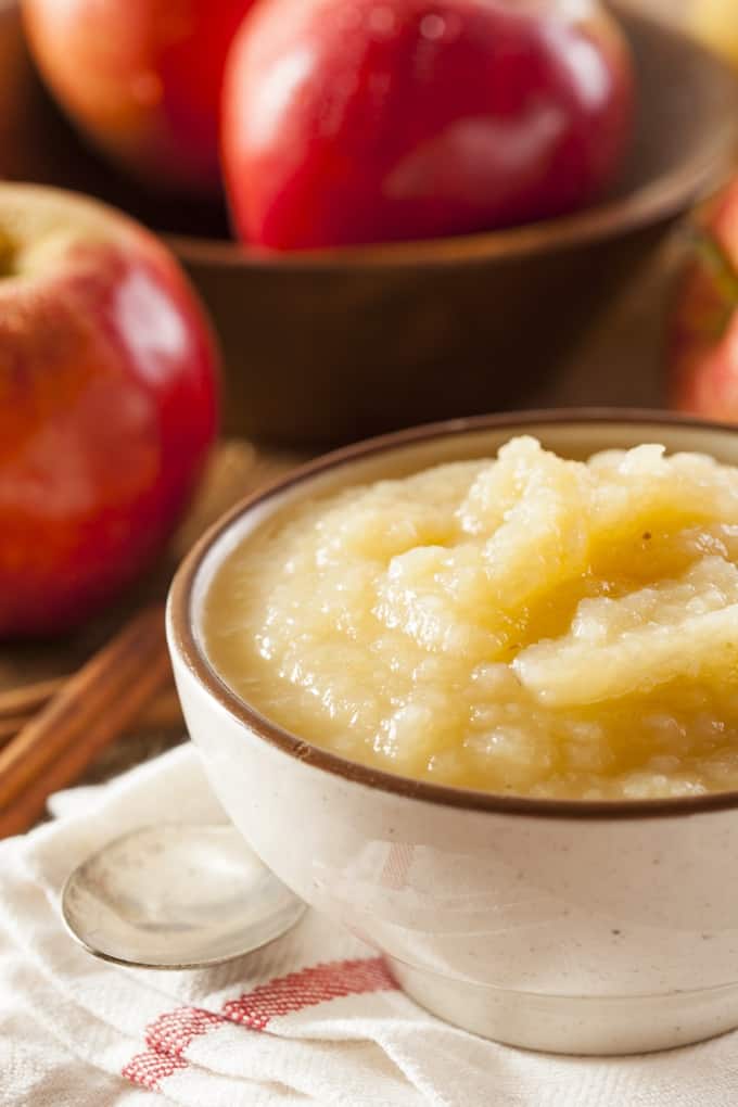 Applesauce in white dish next to spoon and bowl with fresh apples.