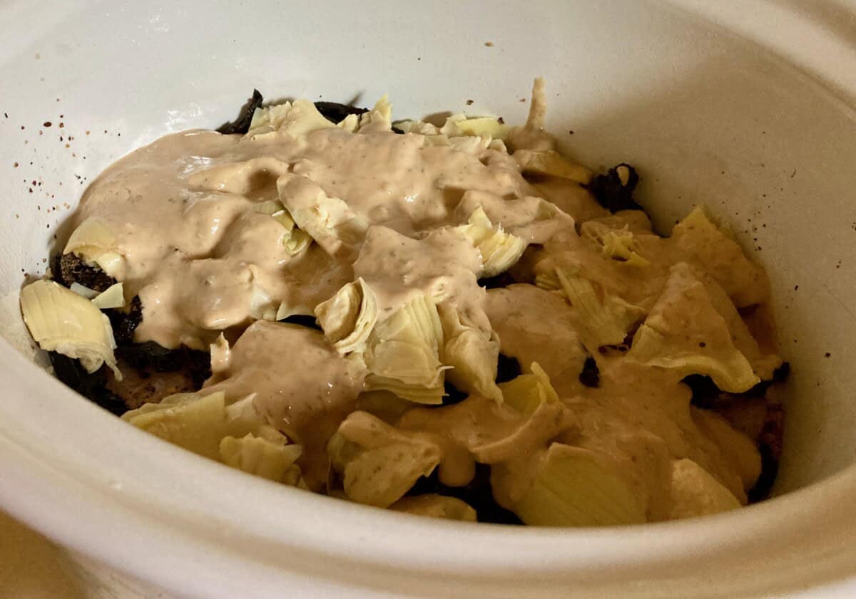 chicken, sun dried tomatoes, quartered artichoke hearts drizzled with dressing in the crock pot ready to cook