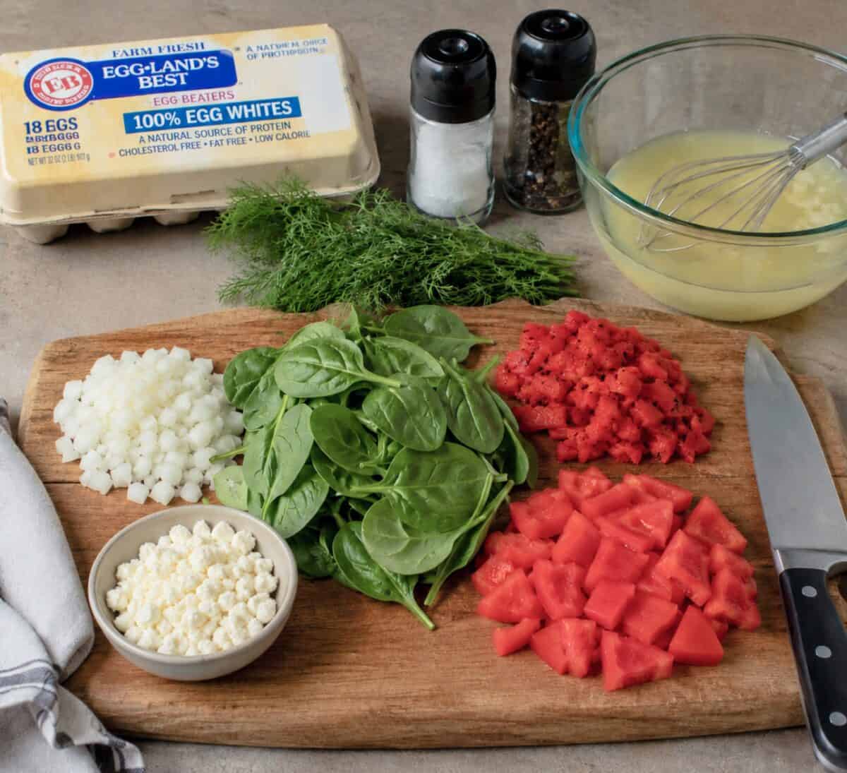 Frittata ingredients including a carton of eggs, fresh spinach, crumbled feta, chopped onion, fresh dill, chopped tomatoes, chopped roasted red peppers and a glass bowl with eggs and whisk.