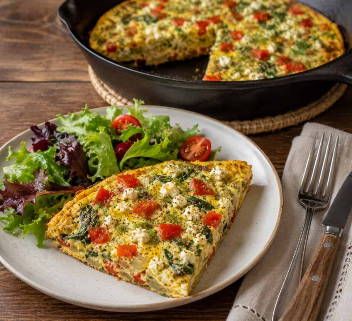 Slice of Greek Frittata on a white plate with a mixed green salad, fork and knife, and skillet with remaining frittata in the background.