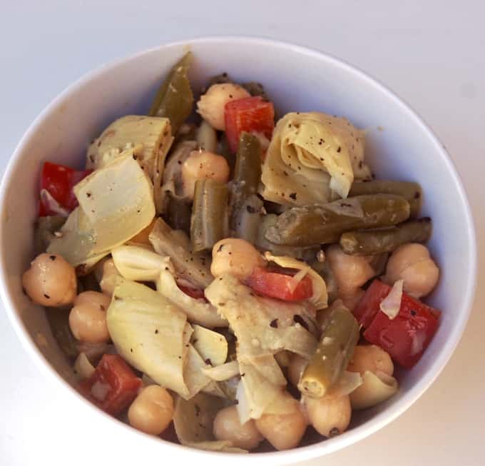 Two bean salad with green beans, chickpeas, artichokes and red peppers in white bowl.