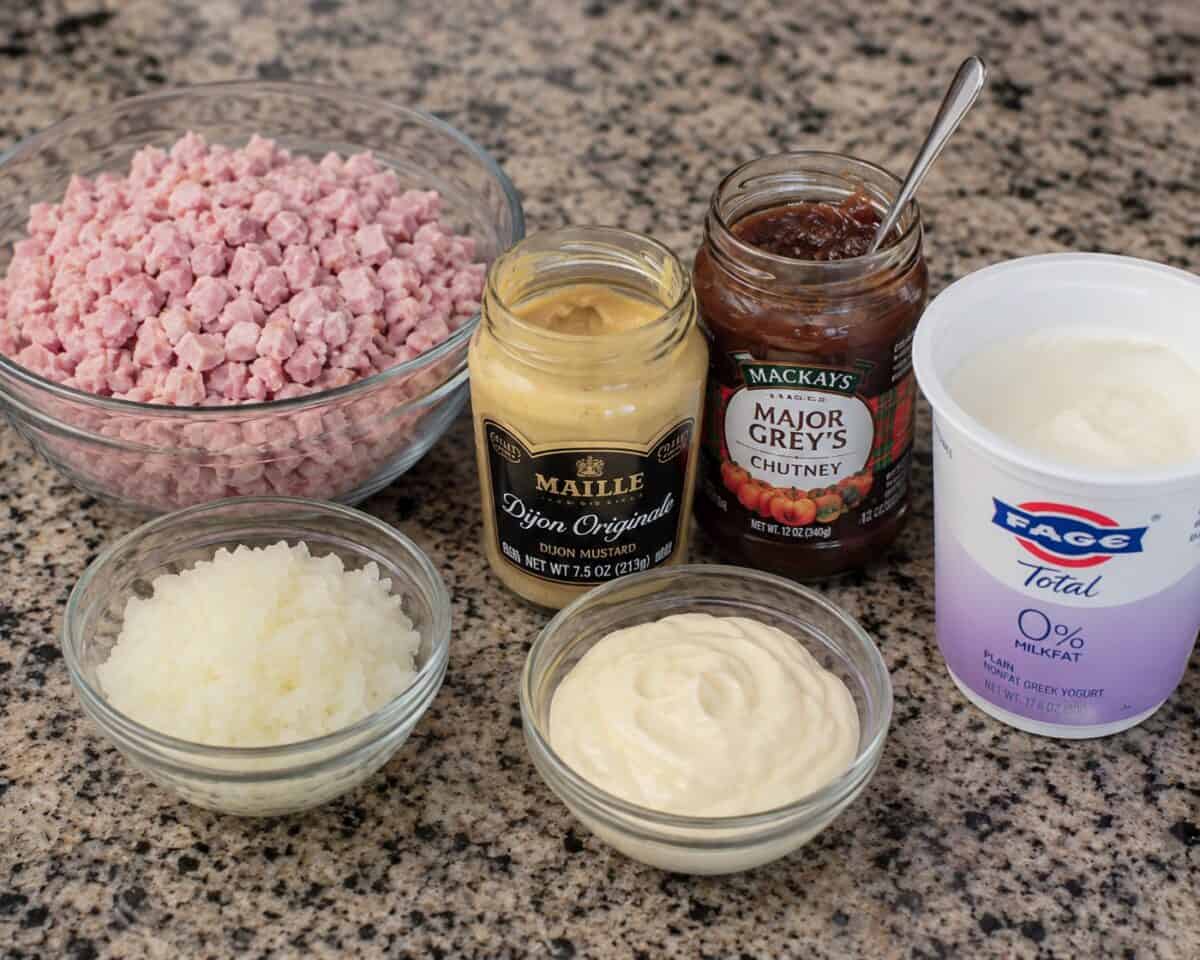 Ingredients including a bowl of chopped cooked ham, grated onion, light, light mayonnaise, a jar of mustard, a jar of chutney and a container of Greek yogurt.