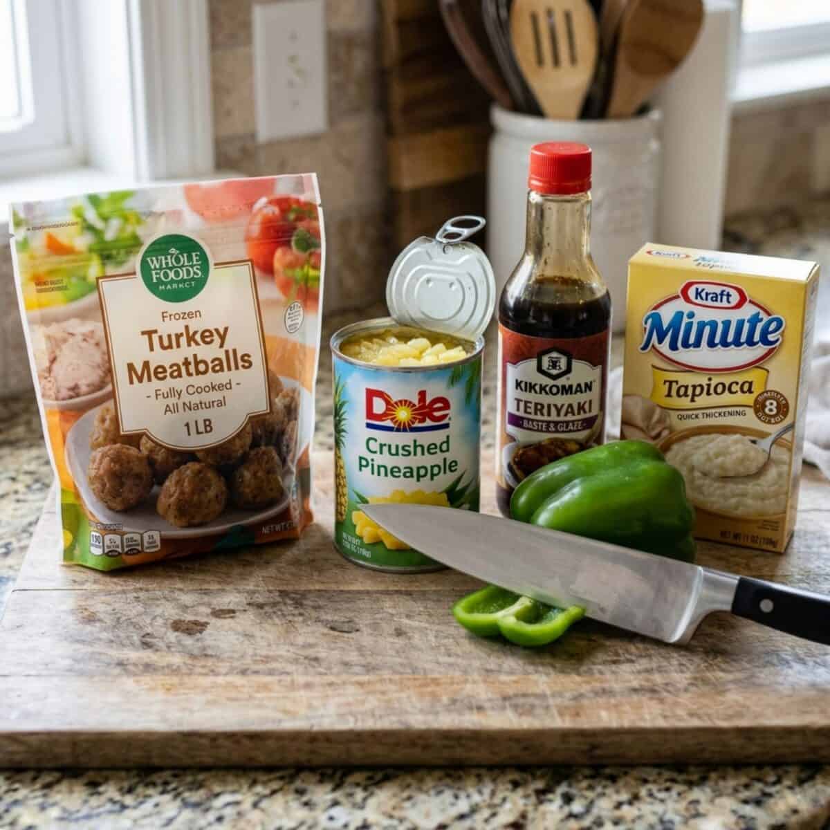 A bag of frozen turkey meatballs, an open can of crushed pineapple, a jar of Kikkoman teriyaki baste and glaze, a box of minute tapioca and a green bell pepper on a wood cutting board with a chef's knife.