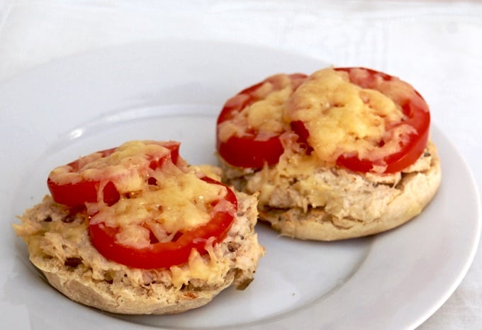 Two english muffin halves topped with tuna salad, sliced tomato and melted cheese on a white plate.