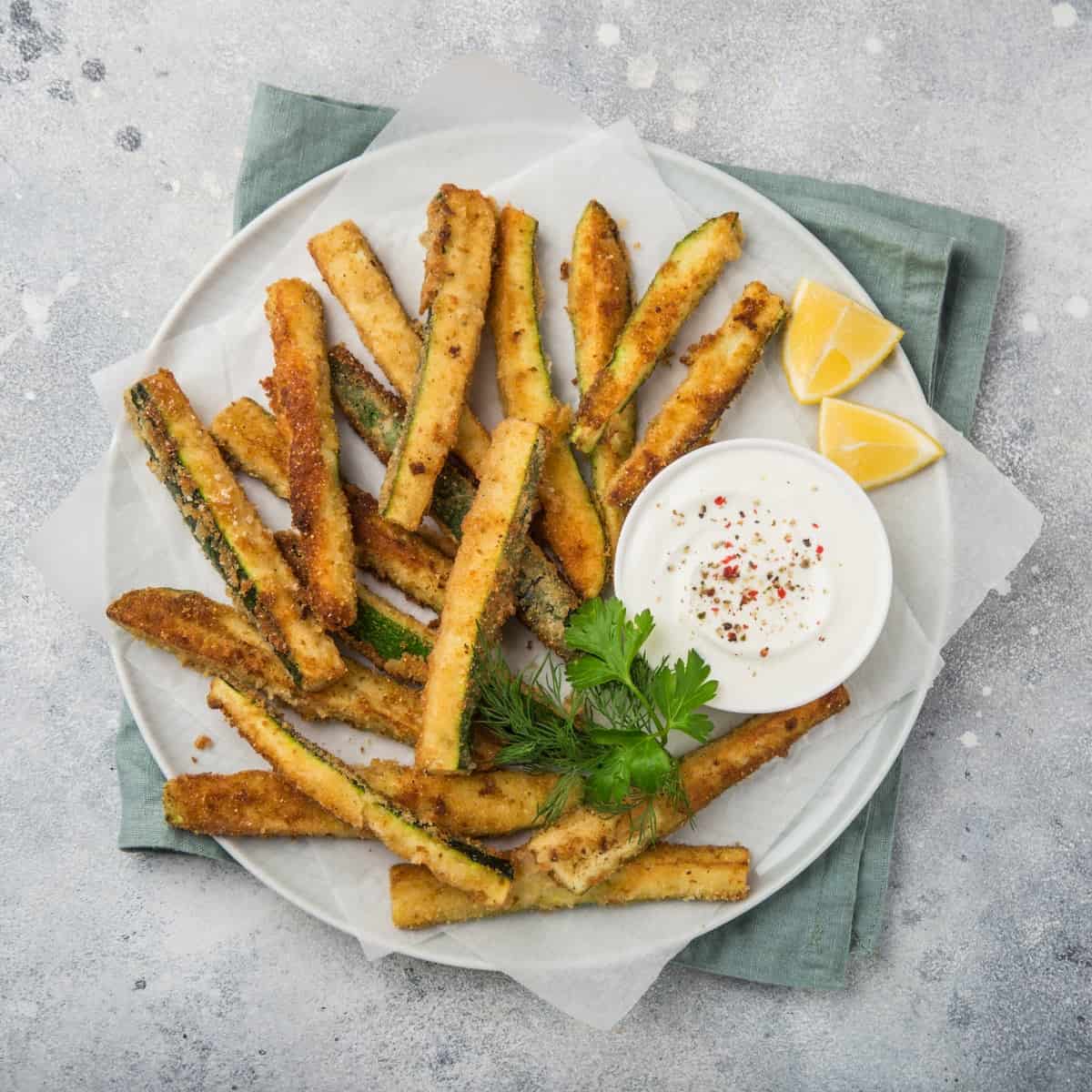 Baked zucchini fries on white plate with Greek yogurt dipping sauce and lemon wedges from above.
