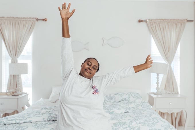 Smiling young African woman sitting alone on her bed with her arms in the air stretching while waking up in the morning