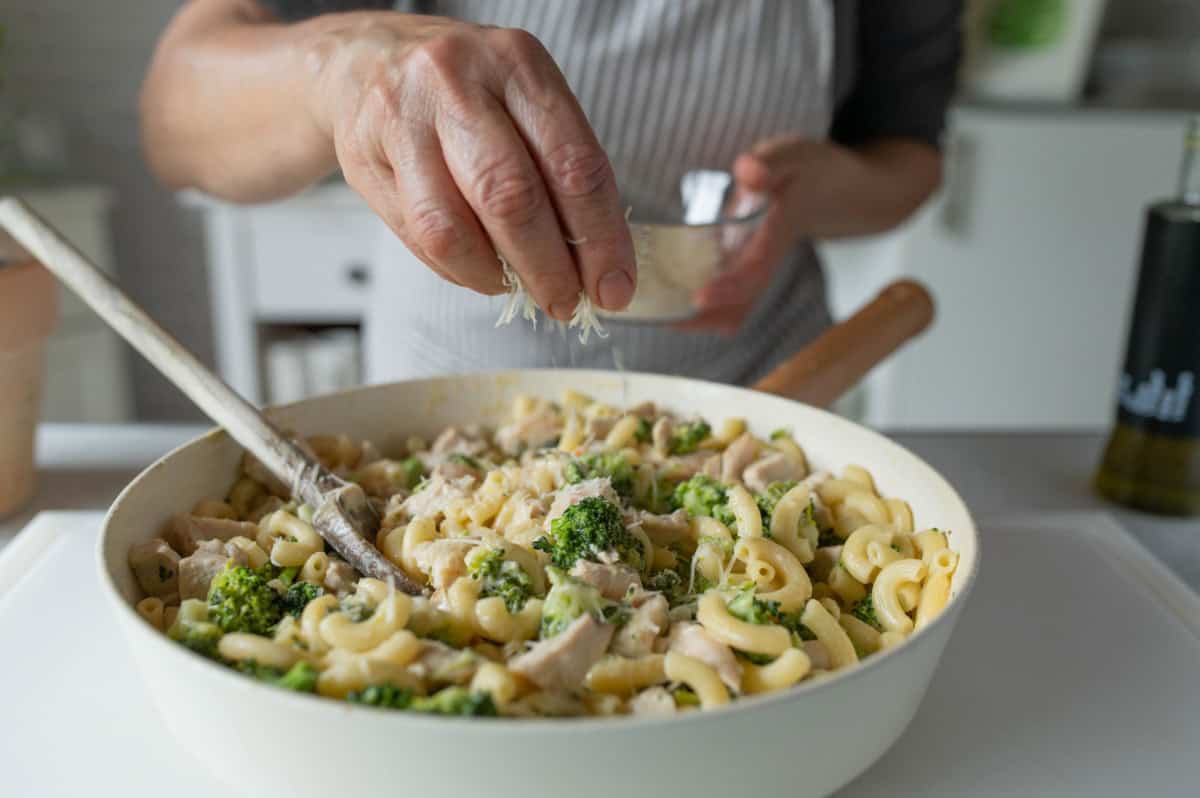 Sprinkling grated Parmesan cheese to skillet with macaroni pasta, broccoli and chicken.