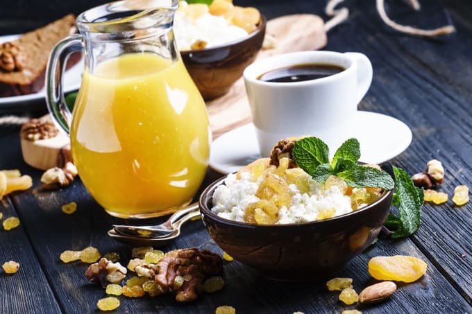 Cottage cheese in a bowl with golden raisins with white cup of coffee and pitcher of orange juice behind.