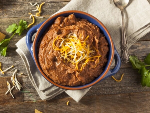Small blue bowl filled with refried bean dip topped with shredded cheese on a wooden table with a linen napkin and spoon