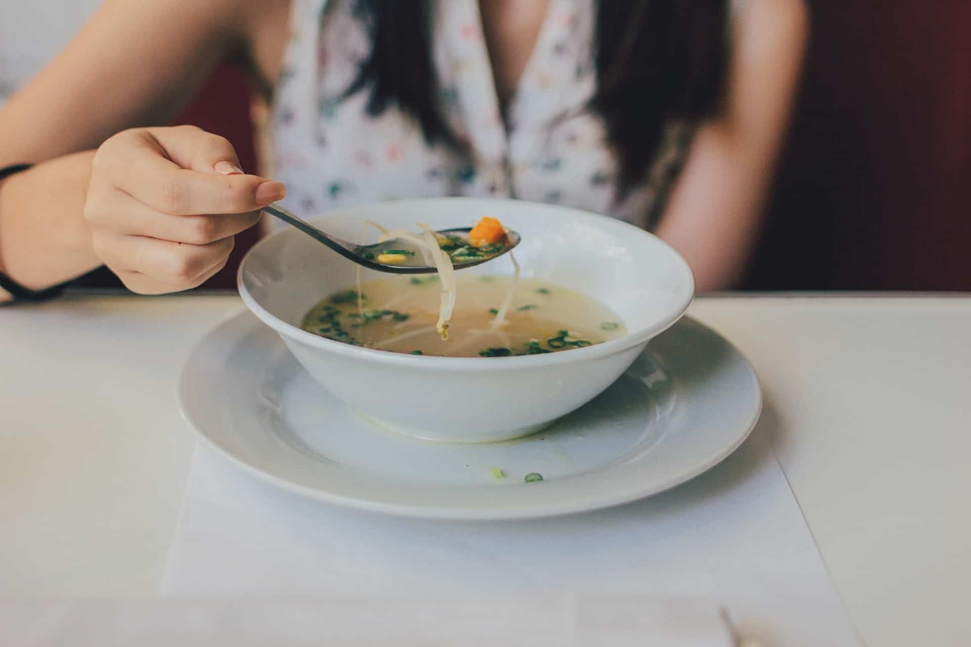 Front shot of woman eating bowl of soup from a white bowl on a white plate.