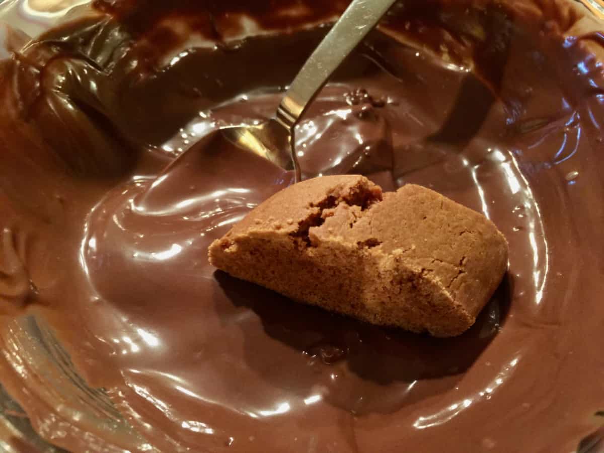 Glass bowl of melted dark chocolate with molasses cookie slice bottom being dipped into it.
