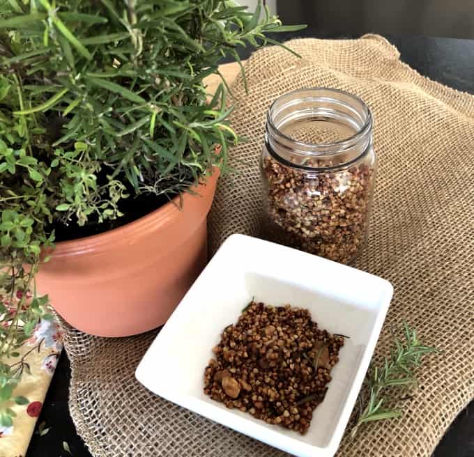 Buckwheat Crunch in a white dish on a burlap sack next to a mason jar with more buckwheat crunch and a potted rosemary plant.