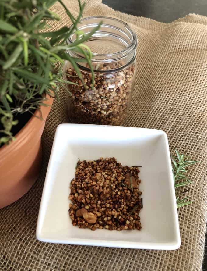 Rosemary Buckwheat Crunch in a square white dish on a piece of burlap next to a mason jar with more rosemary buckwheat crunch and a fresh rosemary plant