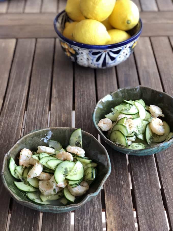 Two bowls with Parisian shrimp and cucumbers on wood table with bowl of lemons in the background.