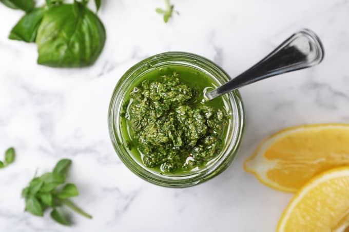Jar of basil pesto from above with spoon, basil leaves and lemon wedges on white marble