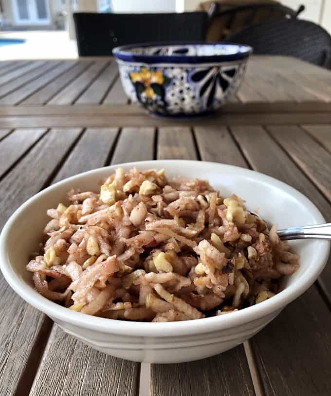 Charoset with shredded apples and chopped walnuts in white bowl with decorative pottery in background.