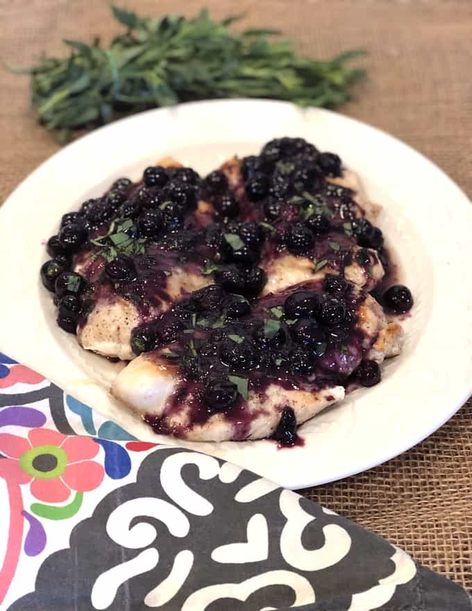 Chicken breasts topped with fresh blueberry sauce on white plate with bunch of fresh tarragon in background.