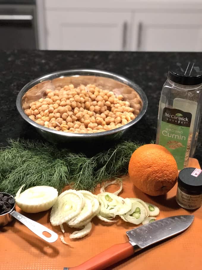 Ingredients including chickpeas in bowl, ground cumin, sliced fennel and an orange.