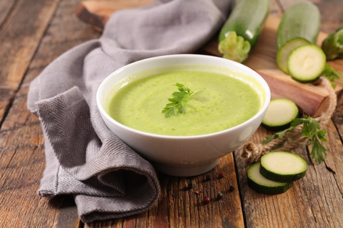 Bowl of creamy zucchini soup on wood table with fresh slices of zucchini.