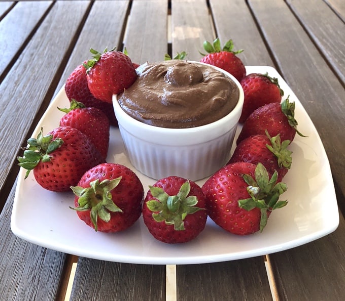 Chocolate brownie batter dessert hummus in small white bowl with plate of fresh strawberries.