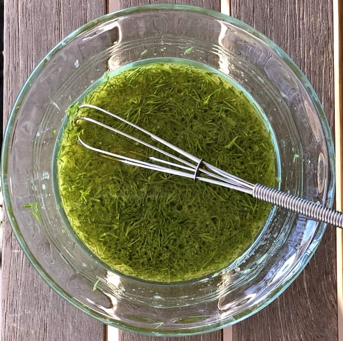 Olive oil, red wine vinegar, Dijon mustard and fresh dill being whisked in small glass bowl.