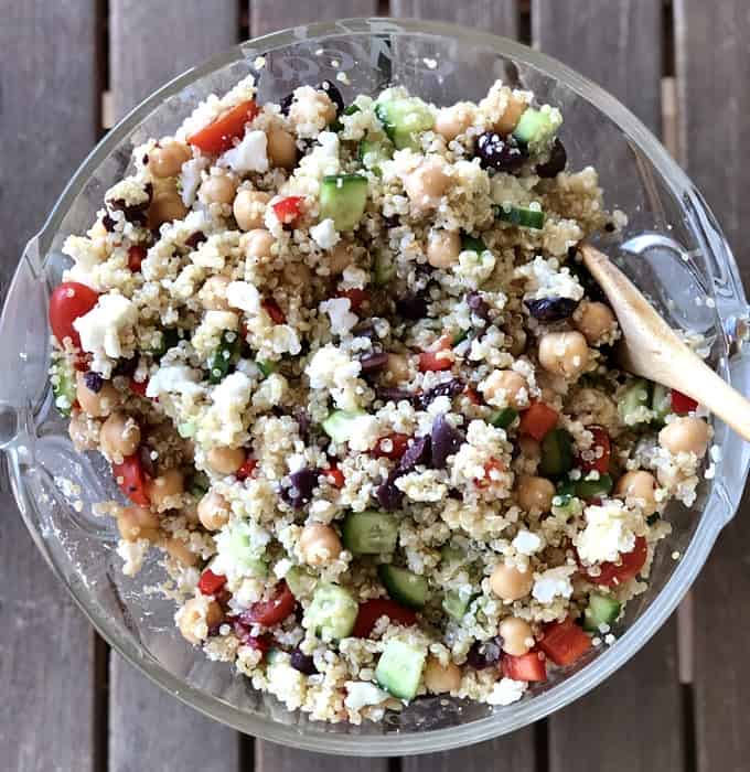 Mixing quinoa, feta cheese, cucumber, red bell pepper, tomatoes and Kalamta olives in glass bowl with wooden spoon.