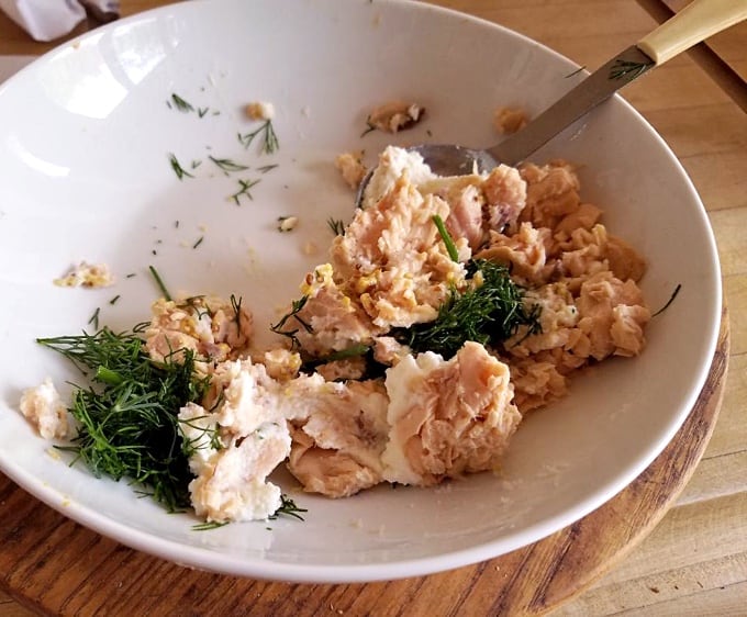 Mixing canned salmon, ricotta cheese, Dijon mustard and chopped dill in white bowl with spoon.
