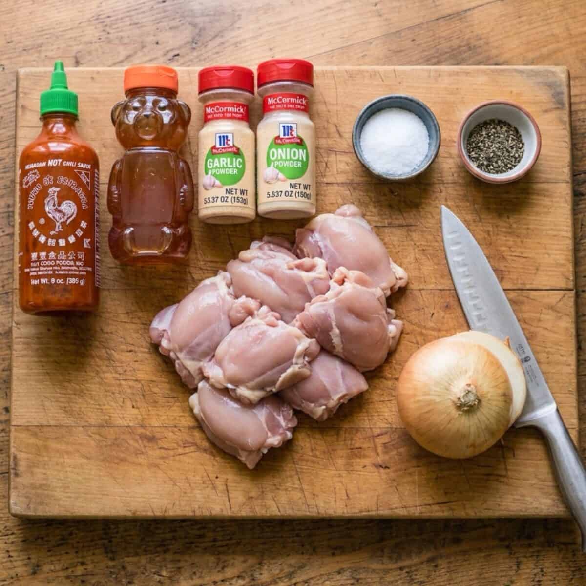 Ingredients including a bottle of Sriracha, bottle of honey, garlic powder, onion powder, salt and pepper, boneless skinless chicken breasts and onion with knife on wood cutting board.