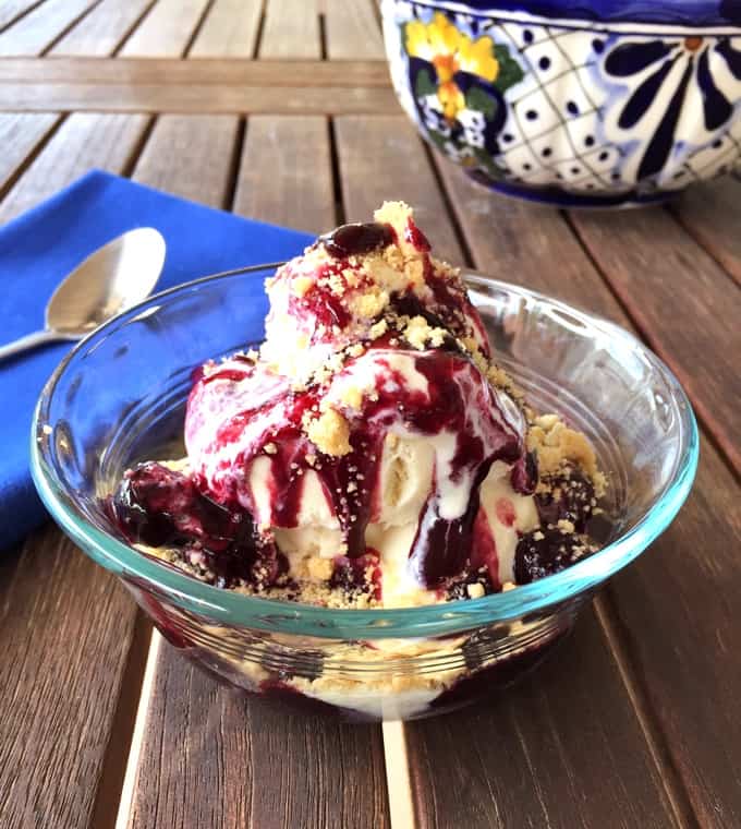 Blueberry pie sundae with crumbled shortbread finger cookies in glass bowl with spoon on wooden table.