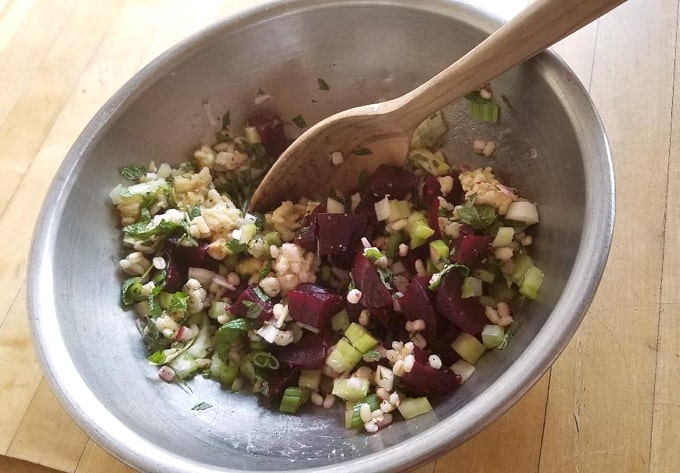 Stirring beets, barley, celery, green onion in stainless mixing bowl with wooden spoon.