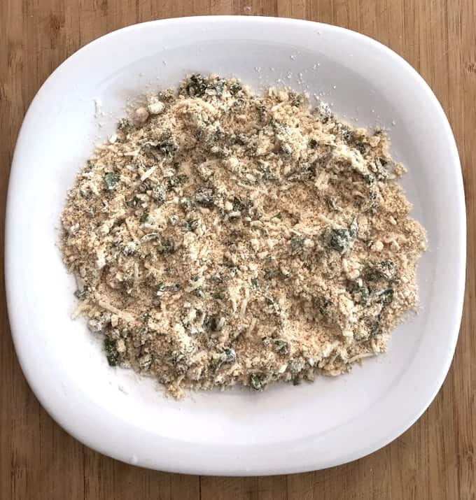 Cashew flour, white flour, grated Parmesan and chopped basil in shallow white plate for coating baked cheese sticks.