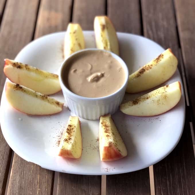 Fresh apple slices topped with cinnamon on white plate with bowl of creamy peanut butter dip.