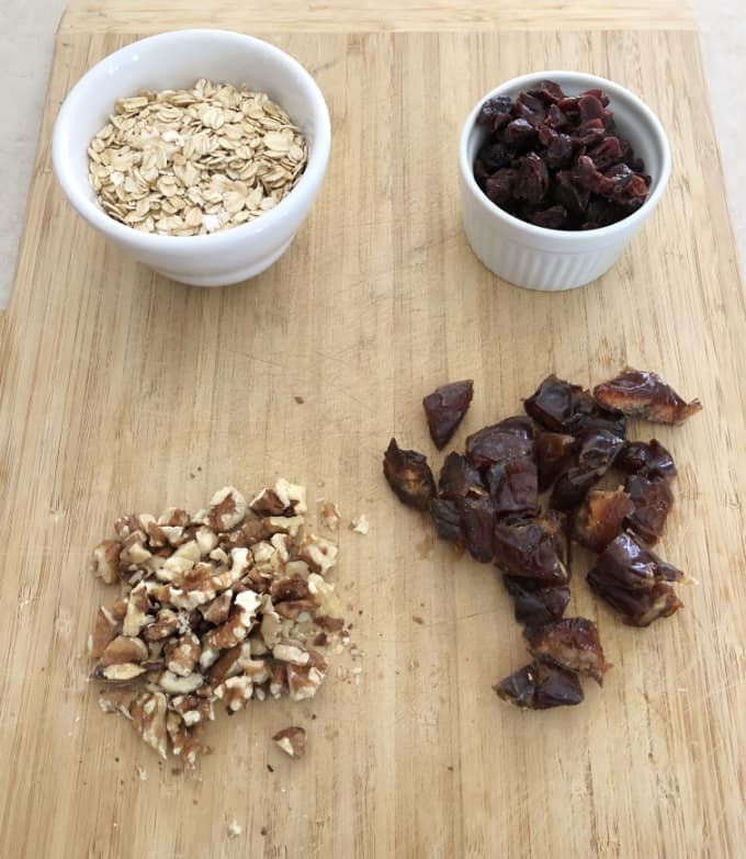 Rolled oats in white bowl, dried cranberries in white bowl, chopped dates and chopped walnuts on bamboo cutting board.